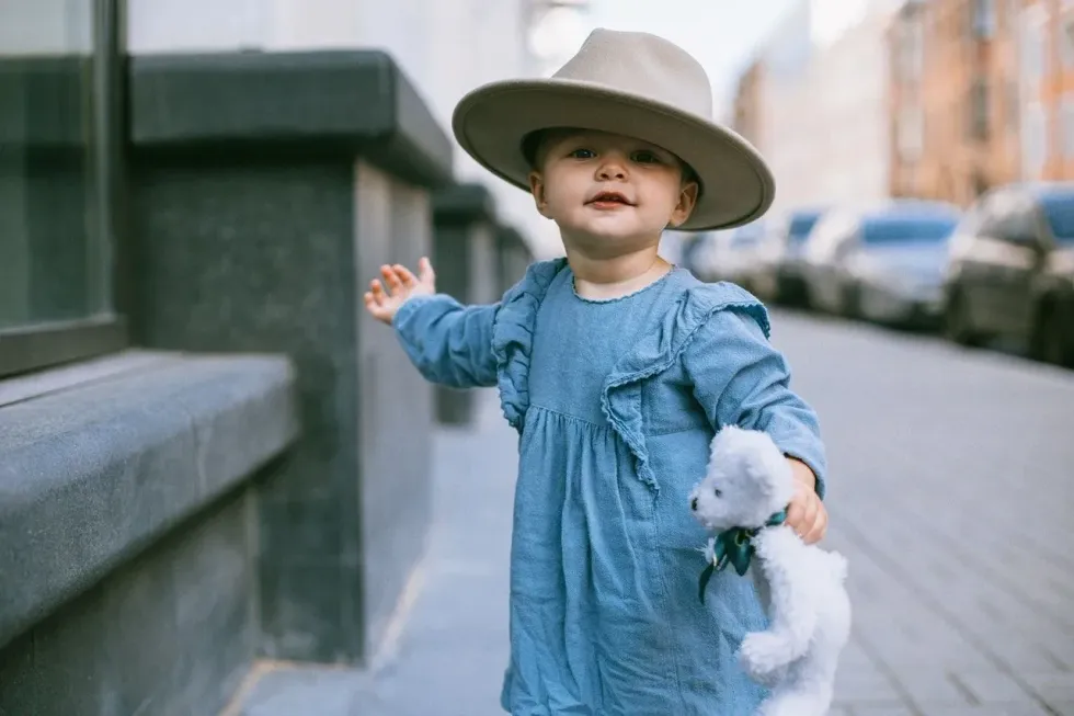 A baby girl in a blue dress and a hat holding a teddy bear