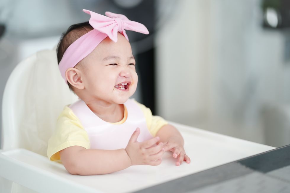 A baby girl wearing a pink hair bow, laughing on a white chair.