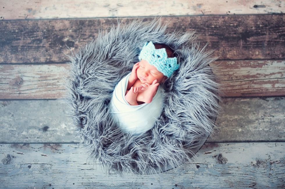 A contented newborn wrapped in white, nestled on a fluffy grey blanket with a blue crown, reminiscent of posh baby boy elegance.