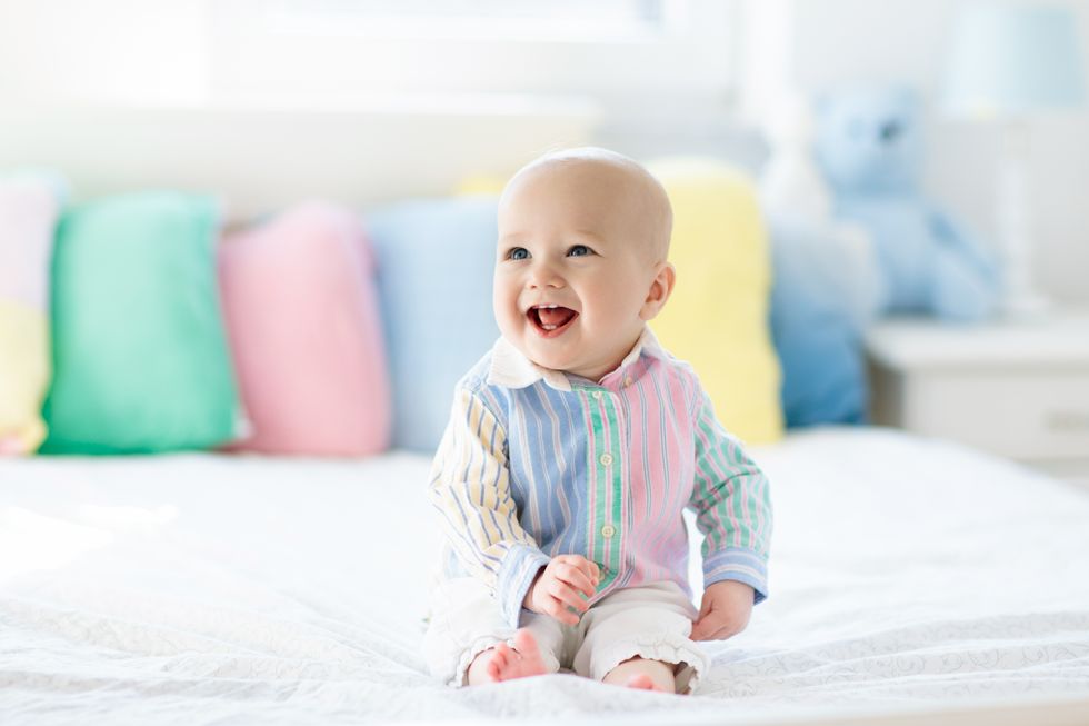 A joyful posh baby with a radiant smile, sitting on a bed in a pastel-striped shirt, with colorful pillows in the background.