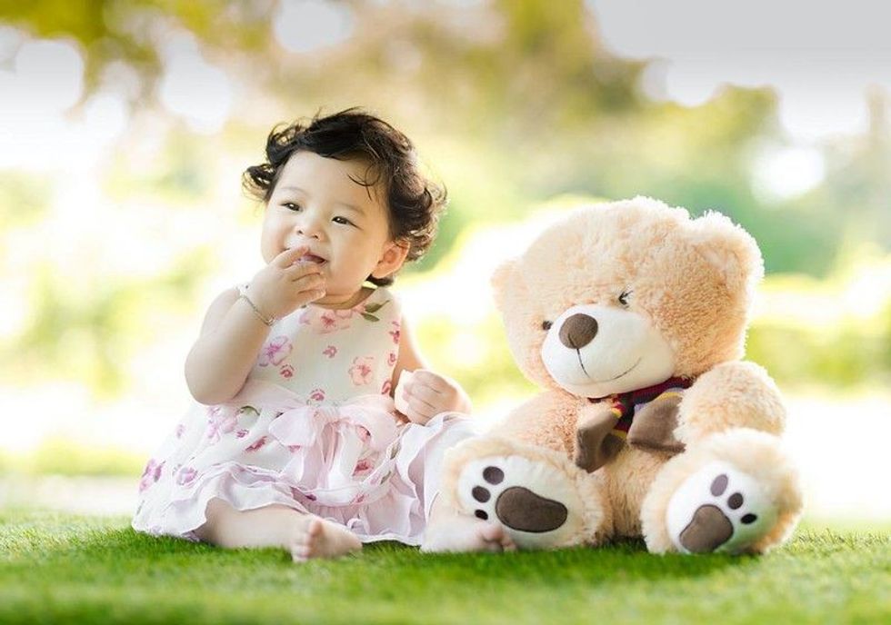 A little girl sitting on grass with a teddy bear beside her.