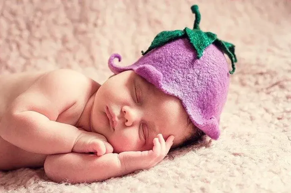 A newborn baby wearing a brinjal hat is sleeping on a pink blanket