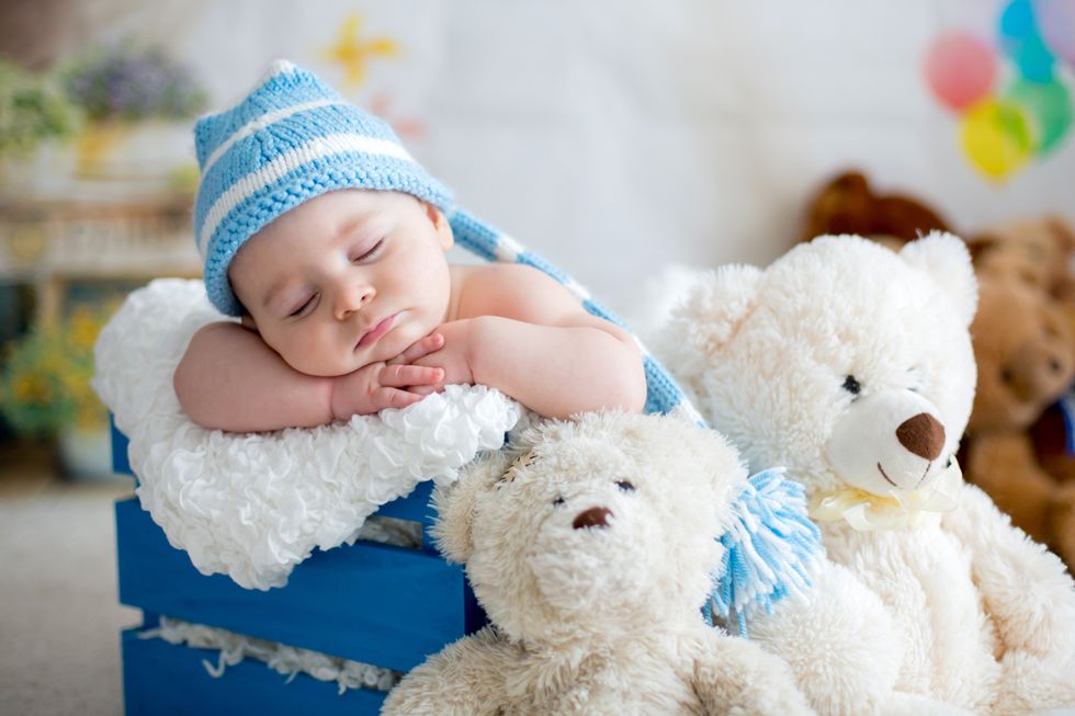 A posh baby boy in a blue striped hat sleeps peacefully on a white fluffy pillow, cradled by stuffed teddy bears.