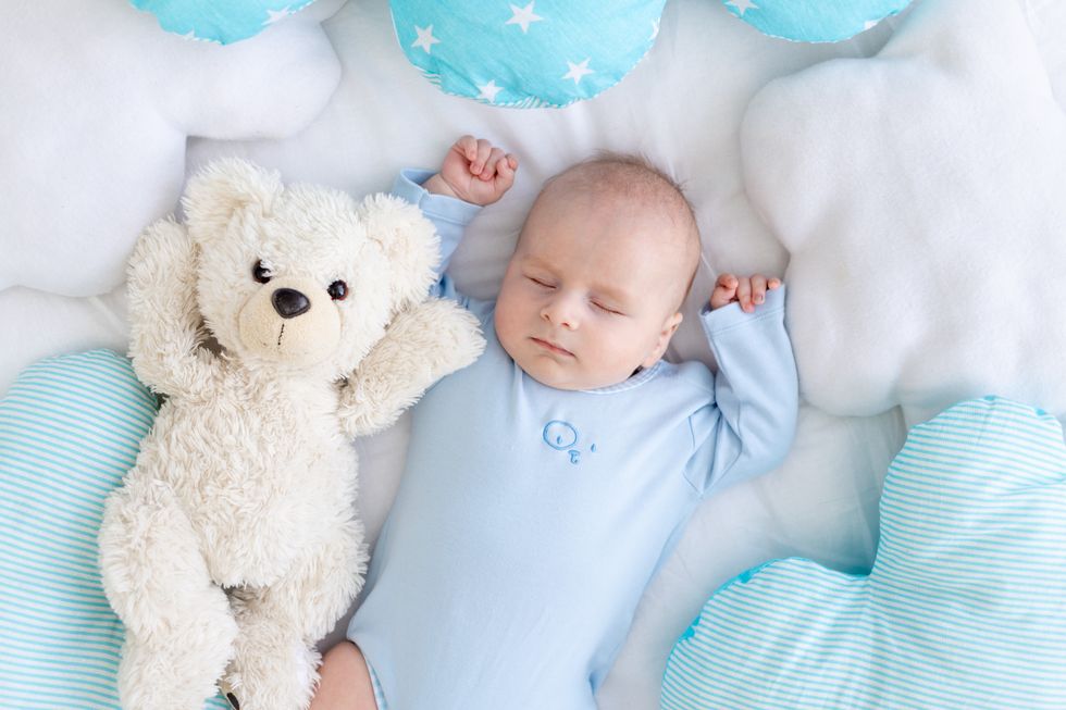 A posh baby in a blue onesie lies comfortably on a cloud-shaped pillow with a cuddly teddy bear and star-patterned cushions around.