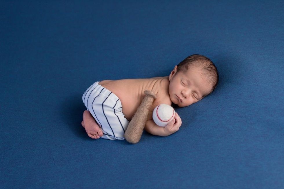 A week old, sleeping, newborn baby boy wearing baseball uniform pants and holding a baseball.