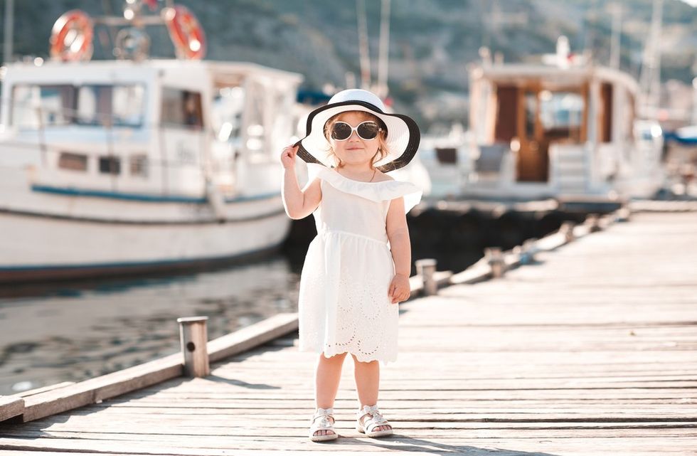 An image of a little girl standing on a deck with white boats parked on water behind her.  She is dressed fashionable in a white dress, a white sandal, a white sunglass and a white hat with black layer at the helm