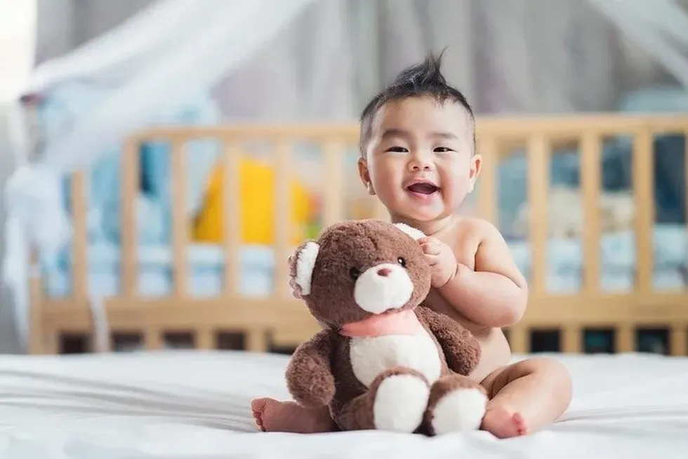 Baby boy sitting on the bed smiling with a teddy bear in arms.