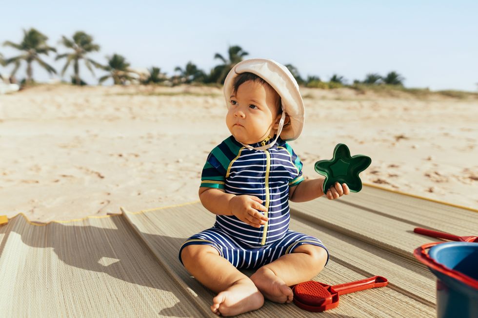 Baby sitting on a mat and playing with toys on a beach.