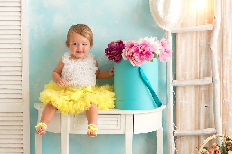 Cute baby girl wearing yellow tulle dress sitting next to flower vase on a table.