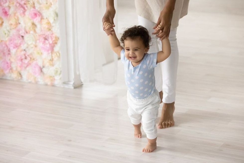 Smiling little biracial toddler infant child make first steps at home holding mom hands.