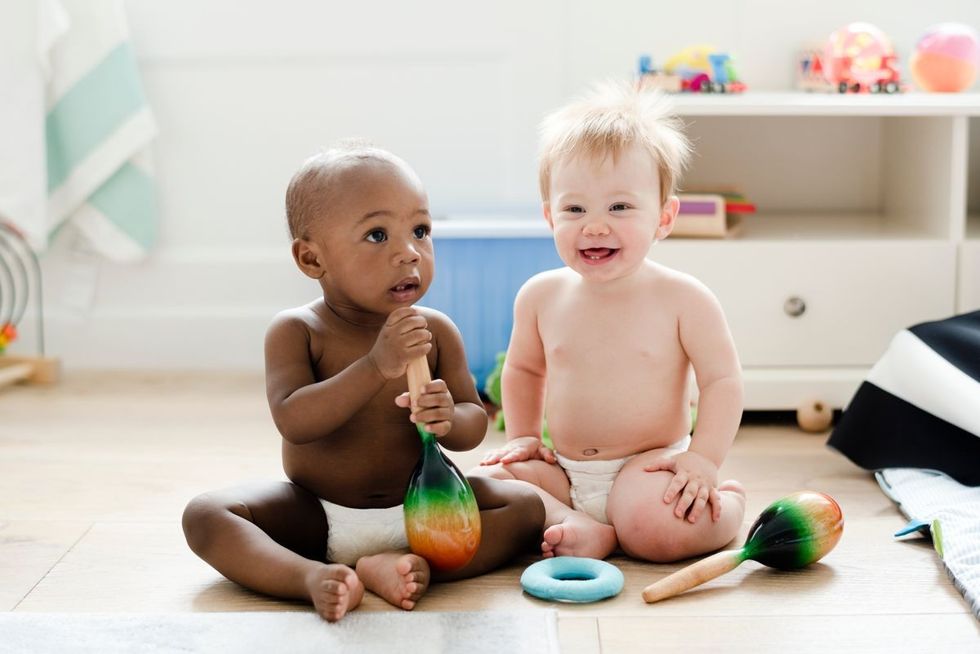 Two baby boys sitting on the floor, one holding a maraca, depicting the joy of a melodic name for boys.