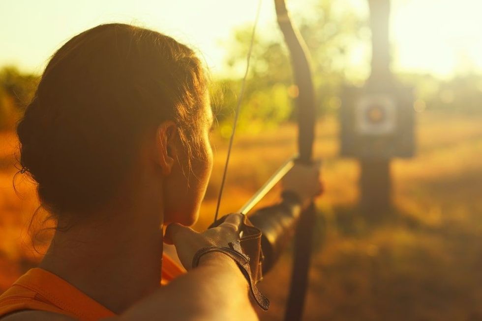 Young female archer shooting with a bow in a field at sunset